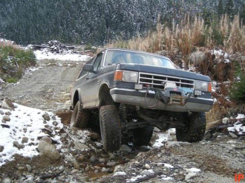 1985 Ford Bronco - A dark colored Ford Bronco is navigating a rocky, snowy trail in a wooded area. The vehicle is equipped with large off-road tires and a win…