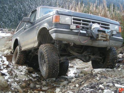 1985 Ford Bronco - A dark-colored, lifted 1980s Ford Bronco is navigating a rocky, muddy trail with snow visible on the ground and trees in the background. Th…