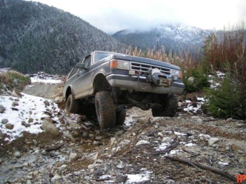 1985 Ford Bronco - A lifted, dark blue 1985 Ford Bronco with a winch on the front bumper navigates a rocky, snow-dusted trail in a mountainous, wooded area.