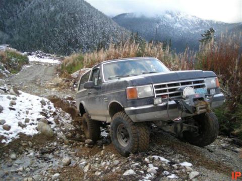 1985 Ford Bronco - A dark-colored Ford Bronco is shown navigating a rocky, off-road trail with patches of snow. The vehicle is equipped with a winch mounted o…