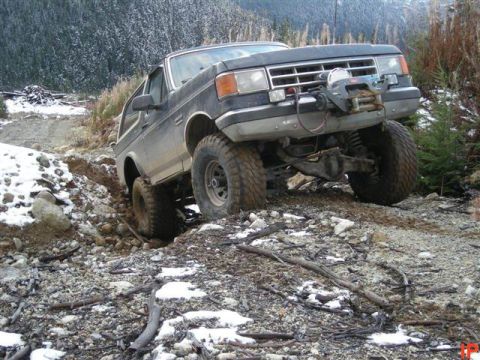 1985 Ford Bronco - A dark colored Ford Bronco is navigating a rocky, off-road trail with snow patches on the ground and trees in the background.