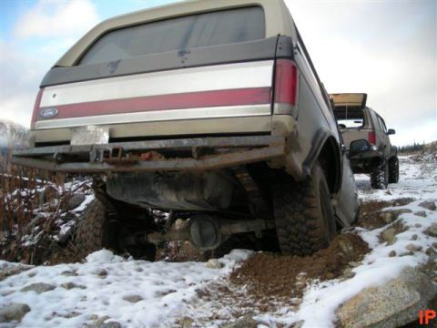 1985 Ford Bronco - The rear of a tan and brown Ford Bronco is stuck in mud and snow on an off-road trail. Another similar SUV is visible behind it.