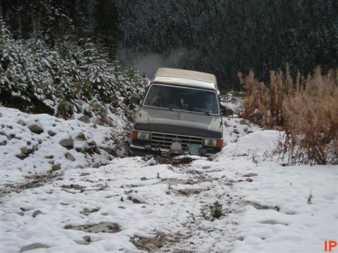 1985 Ford Bronco - A dark-colored SUV is stuck on a snowy, rocky trail surrounded by trees. The vehicle is tilted to the right, with snow covering the ground…