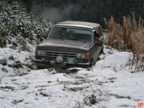 1985 Ford Bronco - A dark colored SUV is stuck in the snow on a dirt road surrounded by trees. The vehicle has a winch mounted on the front bumper and a perso…