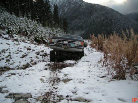 1985 Ford Bronco - A dark-colored SUV with a winch mounted on the front is stuck on a snowy, rocky trail in a mountainous, forested area. The vehicle appears…