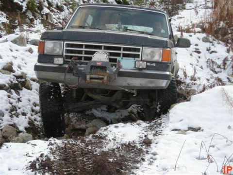 1985 Ford Bronco - A dark colored Ford Bronco is navigating a snowy, rocky trail with a winch mounted on the front bumper. The vehicle is angled towards the v…