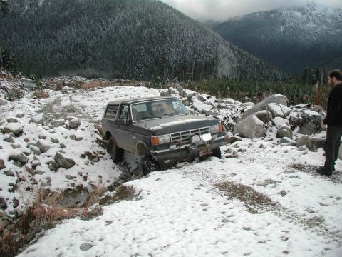 1985 Ford Bronco - A dark-colored Ford Bronco is navigating a rocky, snow-covered trail with a winch mounted on the front bumper. The vehicle is surrounded by…