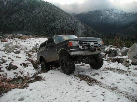 1985 Ford Bronco - A lifted, dark-colored Ford Bronco is navigating a snowy, rocky trail in a mountainous, forested area. The vehicle has large off-road tires…
