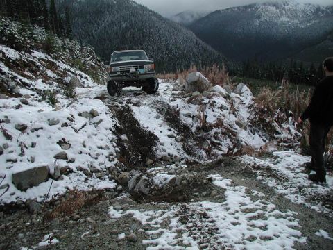 1985 Ford Bronco - A dark-colored 1980s Ford Bronco is navigating a snowy, rocky mountain trail. The vehicle is angled towards the viewer with its front right…