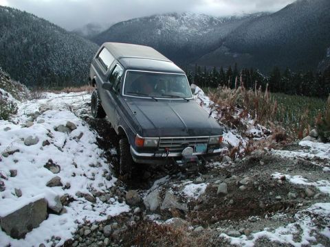 1985 Ford Bronco - A dark-colored Ford Bronco is navigating a snowy, rocky trail with steep, forested mountains in the background. The vehicle appears to be e…