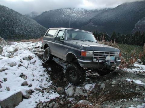 1985 Ford Bronco - A dark blue Ford Bronco is navigating a snowy, rocky trail in a mountainous, forested area. The vehicle has a winch mounted on the front bu…
