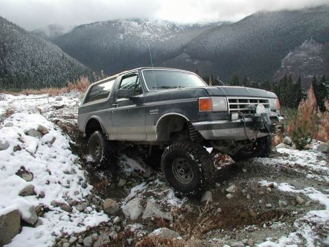 1985 Ford Bronco - A dark blue Ford Bronco is shown navigating a rocky, snow-covered incline in a mountainous, wooded area. The vehicle features large off-roa…
