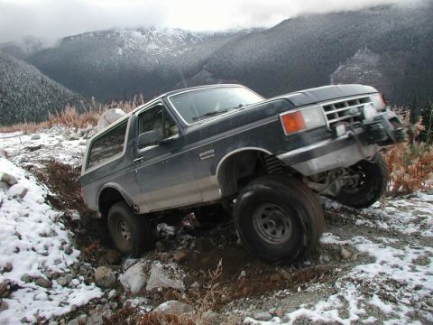 1985 Ford Bronco - A dark blue Ford Bronco is navigating a steep, muddy, and rocky incline with patches of snow. The vehicle is angled with its front wheels t…