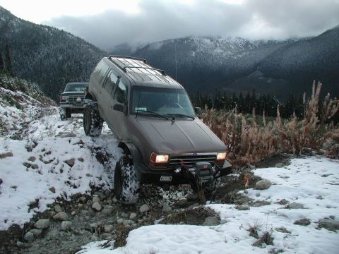 1985 Ford Bronco - A brown SUV with large tires navigates a rocky, snow-covered trail with a winch mounted on the front bumper. Another older model SUV is vis…