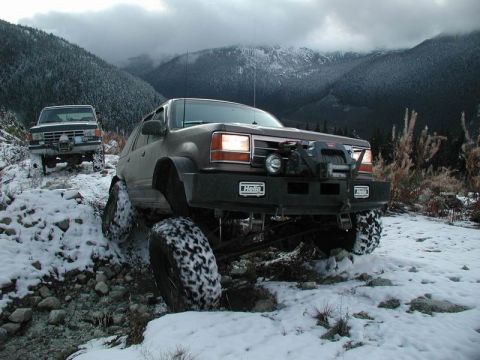 1985 Ford Bronco - Two modified off-road SUVs navigate a rocky, snow-covered mountain trail under a cloudy sky. The front SUV is a dark color with large tires…