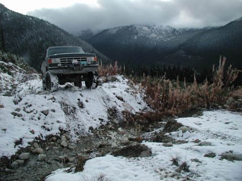 1985 Ford Bronco - A lifted, dark-colored Ford Bronco is parked on a snowy, rocky trail in a mountainous, forested area. The sky is overcast with dramatic clo…
