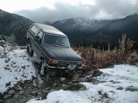 1985 Ford Bronco - A dark-colored Ford Bronco is navigating a snowy, rocky mountain trail with steep inclines and dense evergreen trees in the background.