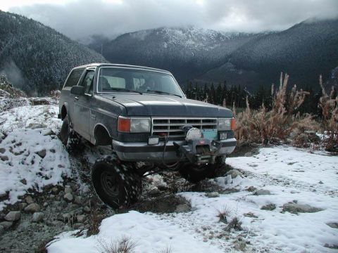 1985 Ford Bronco - A dark colored Ford Bronco is navigating a rocky, snow-covered mountain trail. The vehicle features a winch mounted on the front bumper.