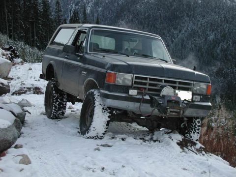 1985 Ford Bronco - A dark blue Ford Bronco is parked on a snow-covered dirt road with large off-road tires. The vehicle has a winch mounted on the front bumpe…