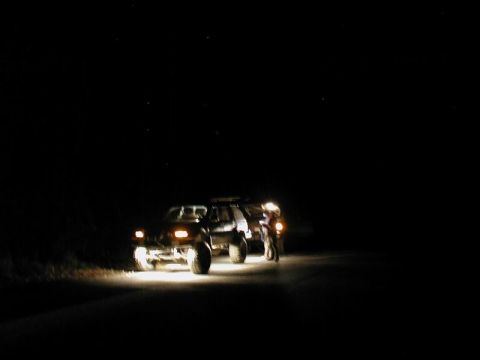 1985 Ford Bronco - Two people stand next to a dark-colored SUV, possibly a Ford Bronco, on a dark road at night. The vehicle's headlights illuminate the road…