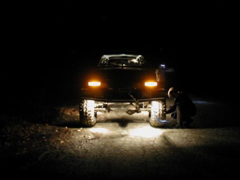 1985 Ford Bronco - A person is airing up the tire of a dark-colored SUV at night, illuminated by the vehicle's headlights and a flashlight.