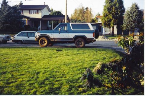1985 Ford Bronco - A two-tone blue and white Ford Bronco is parked on a residential street next to a light-colored sedan. The Bronco has off-road tires and a…