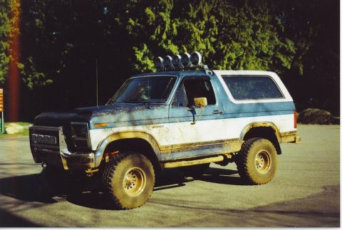 1985 Ford Bronco - A muddy, two-tone blue and white 1985 Ford Bronco with off-road tires and a light bar on the roof is parked on asphalt. The vehicle is view…