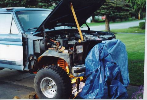 1985 Ford Bronco - The front end of a blue and white Ford Bronco is shown with the hood open, revealing the engine bay. The vehicle is parked outdoors on a pa…