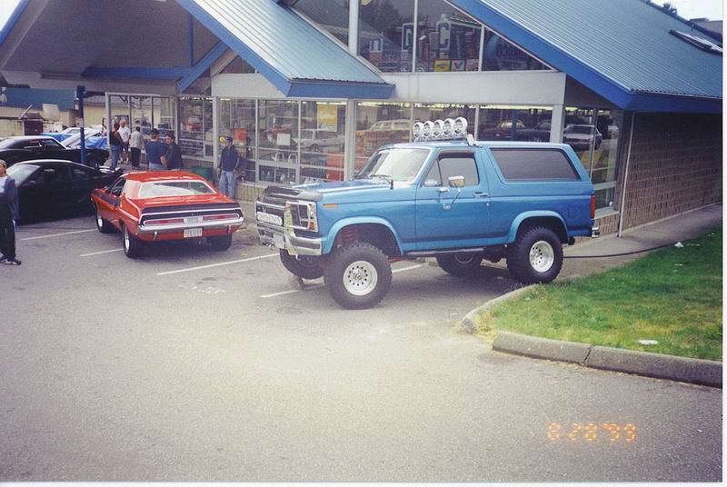 Lifted blue Ford Bronco with roof lights parked next to a red classic car in front of a store.