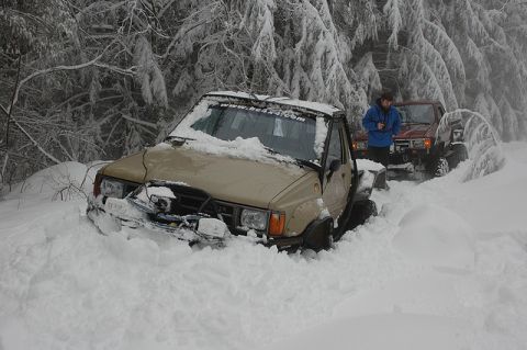 Two off-road trucks stuck in deep snow on a wooded trail.