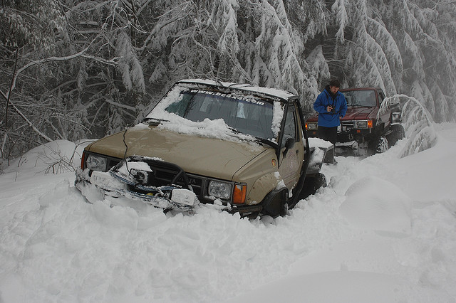 Two off-road trucks stuck in deep snow on a wooded trail.
