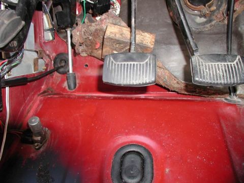 1990 Ford F-250 - The interior driver's side footwell of a red vehicle shows the clutch and brake pedals. Wires and a rubber grommet are also visible.