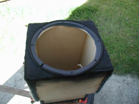 1990 Ford F-250 - An empty, black carpeted subwoofer box with a circular opening is shown from a high angle, resting outdoors on a paved surface with grass i…