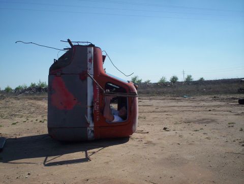 1990 Ford Bronco - A lone truck cab rests on its side in a junkyard, its faded paint and visible company text hinting at a past life.
