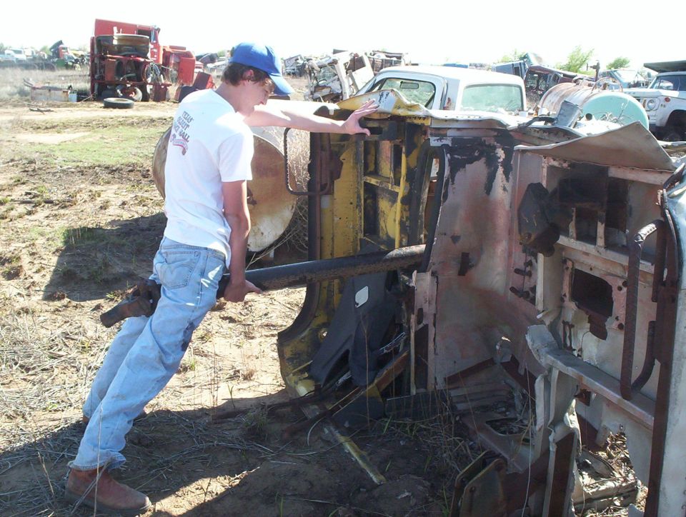 Junkyard scene with overturned vehicle chassis and a person. Other vehicles and parts are visible in the background