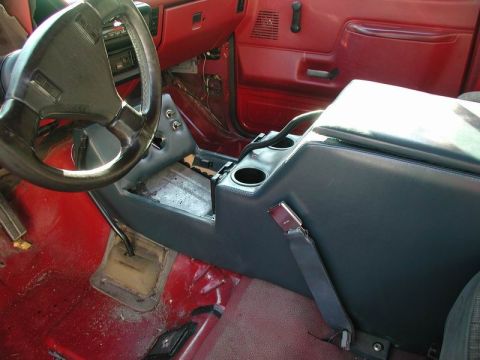 1990 Ford F-250 - The interior of a red Ford F-250 truck shows a dark gray center console with cup holders and a seatbelt. The steering wheel and floor shift…