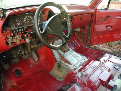 1990 Ford F-250 - The interior of a red 1990 Ford F-250 with the floor stripped down to the metal, revealing the steering wheel, dashboard, pedals, and trans…
