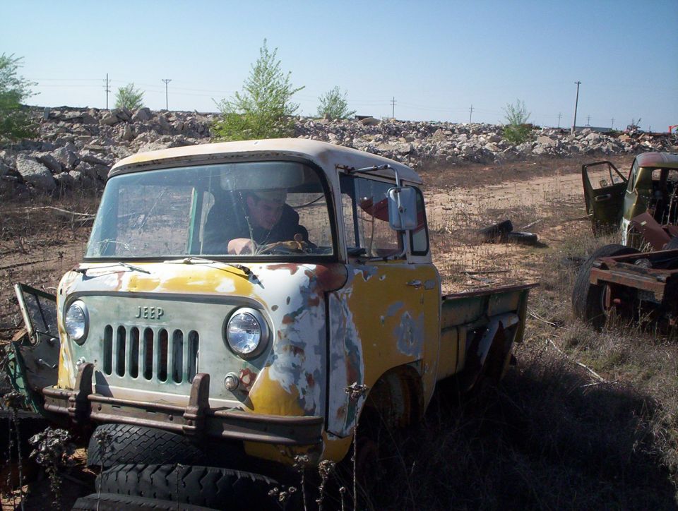 Old Jeep FC-150 pickup truck with a person inside, parked in a junkyard with other vehicles