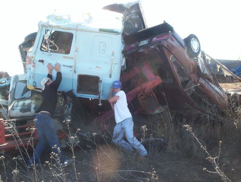 1990 Ford Bronco - Two men attempt to move a wrecked maroon car that is precariously balanced on a light blue truck cab.