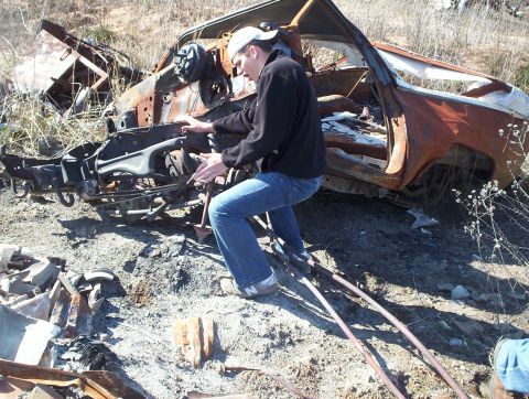 1990 Ford Bronco - A person inspects the heavily damaged and burnt remains of a vehicle, likely at a junkyard.
