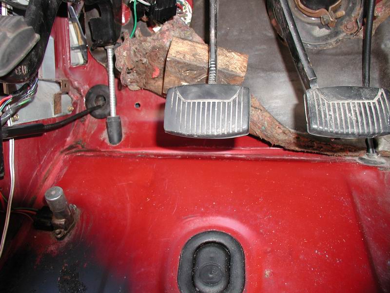 Interior driver's side footwell of a red vehicle showing clutch and brake pedals and wiring.