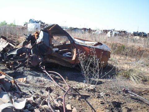 1990 Ford Bronco - Junkyard scene with a heavily damaged and rusted vehicle chassis under a clear sky.