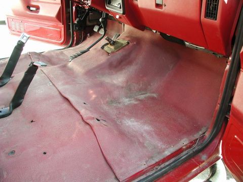1990 Ford F-250 - The interior of a red Ford F-250 pickup truck shows the driver's side floorboard and lower dashboard. The floor is covered in a red rubber…
