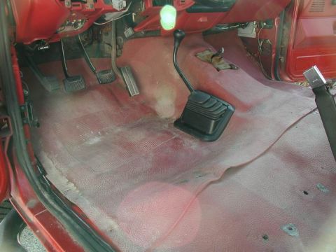 1990 Ford F-250 - The interior of a red truck shows the driver's side floor with pedals, a manual transmission shifter, and worn red carpet. The dashboard an…