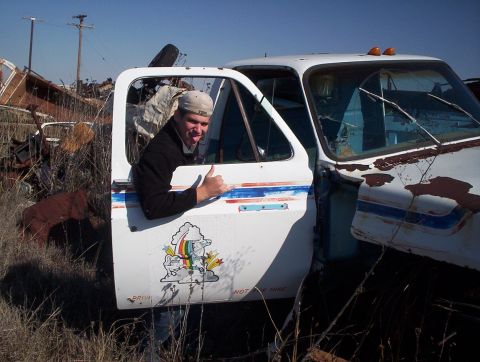 1990 Ford Bronco - A man poses in the open door of a dilapidated pickup truck surrounded by scrap metal and dry grass.