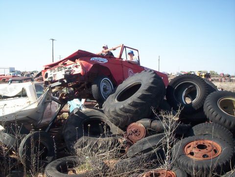 1990 Ford Bronco - A wrecked red Ford Bronco is stacked with tires in a junkyard, with two people inside.