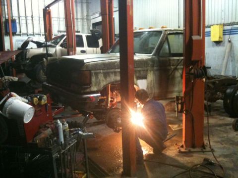 1990 Ford F-250 - A person in a blue jumpsuit is welding underneath the front end of a weathered, older model pickup truck, which is raised on a lift in a ga…
