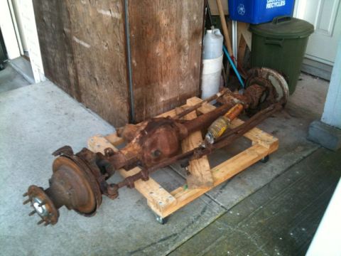 1990 Ford F-250 - A rusty front axle assembly with brake drums and shocks is mounted on a wooden pallet with wheels. The assembly appears to be from a truck,…