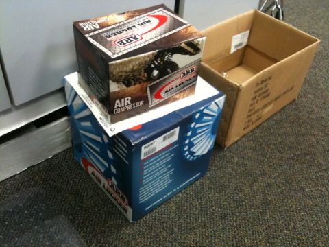 1990 Ford F-250 - Two boxes of ARB air compressors are stacked on top of each other, with an open cardboard box to the right. The boxes are on a carpeted flo…