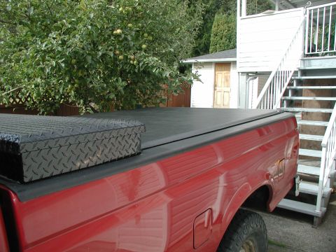 1990 Ford F-250 - The rear passenger side of a red Ford F-250 pickup truck is shown with a black diamond plate toolbox on the bed cover. A white house with a…
